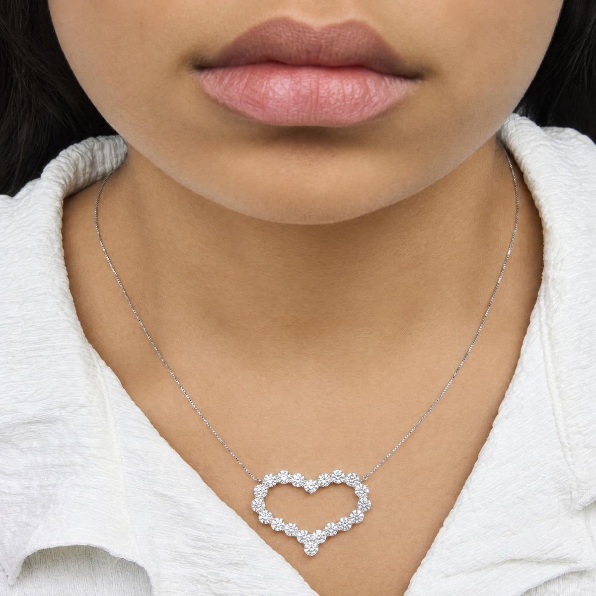 Close-up front view of a model wearing a white metal necklace with a heart-shaped pendant outlined in round white gemstones in prong settings.