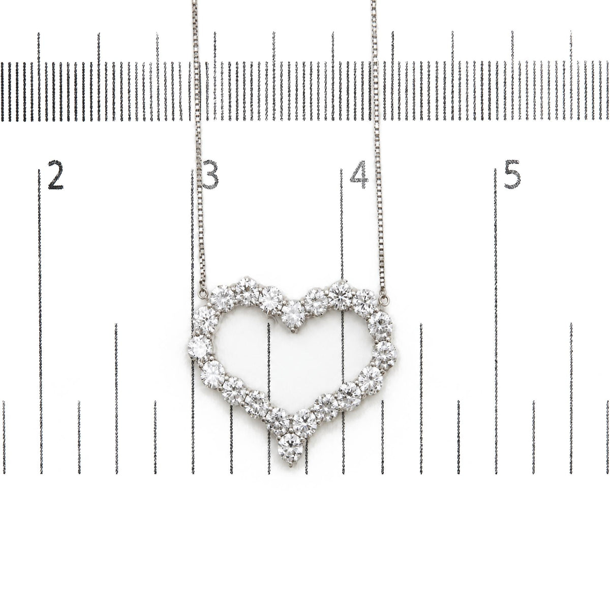 Close-up front view of a white necklace with a heart-shaped pendant made of round white gemstones in prong settings, shown on a measurement scale.