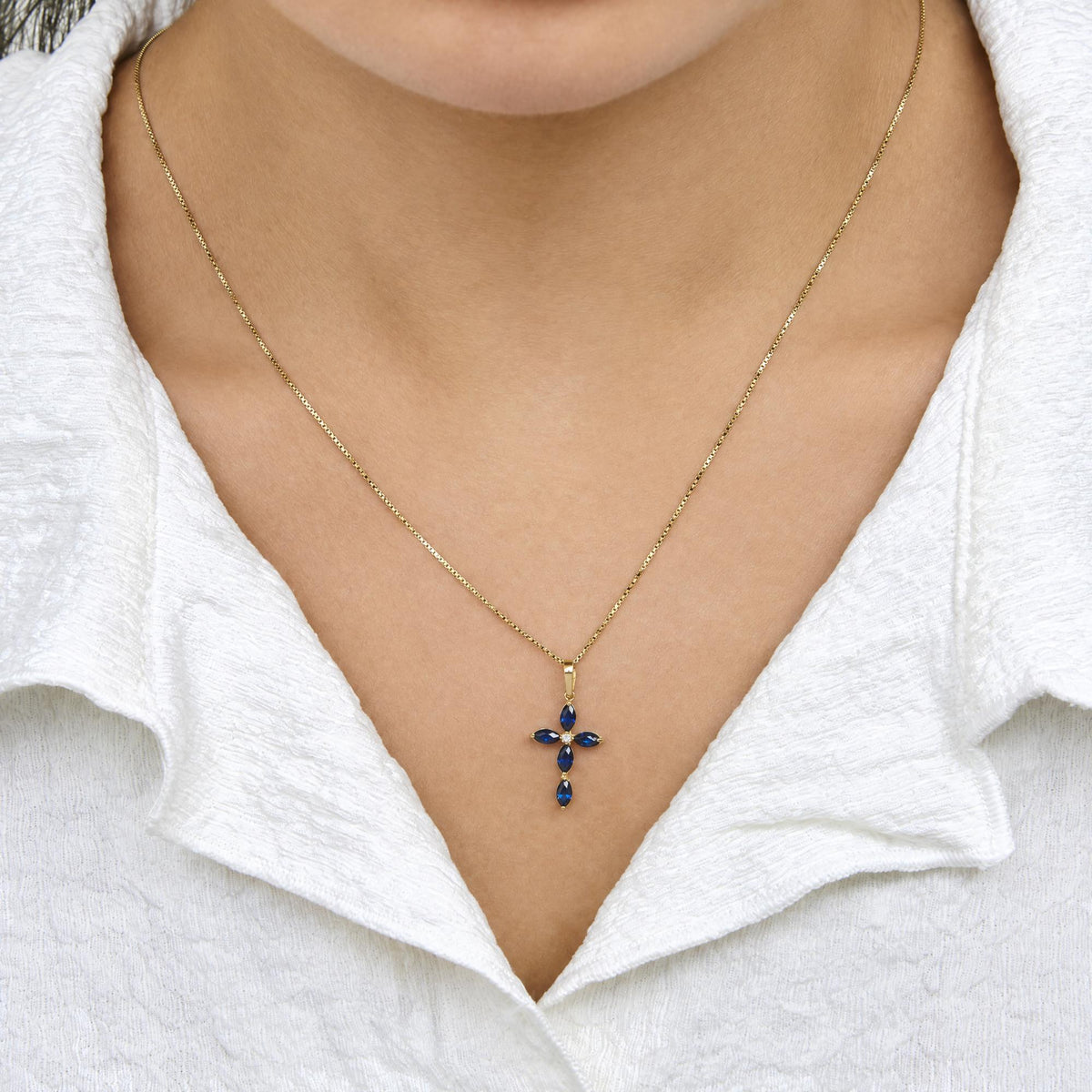 Close-up front view of a model wearing a yellow metal necklace with a cross-shaped pendant featuring blue marquise-shaped gemstones and a single white round gemstone in the center.