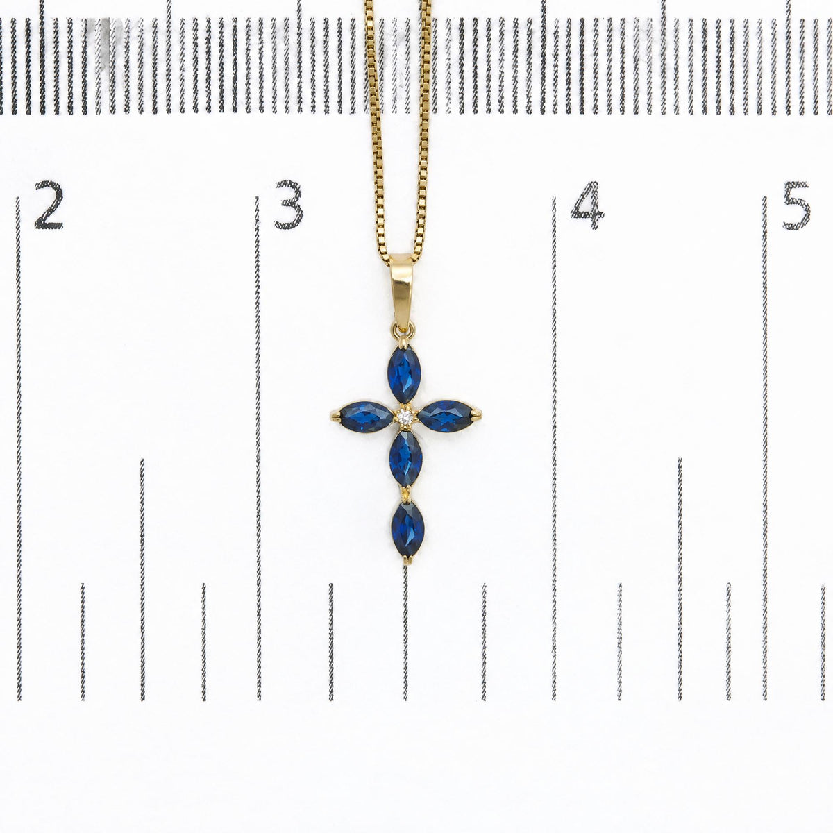 Close-up front view of a yellow metal necklace pendant shown on a scale, featuring a cross shape design with marquise-shaped blue gemstones and one round white gemstone in the center, all with prong settings.