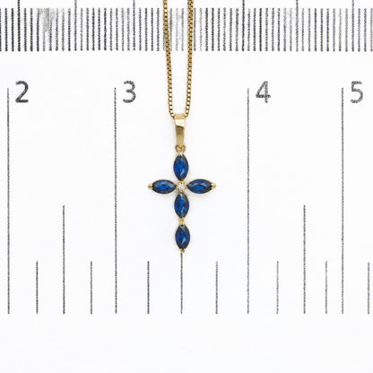 Close-up front view of a yellow metal necklace pendant shown on a scale, featuring a cross shape design with marquise-shaped blue gemstones and one round white gemstone in the center, all with prong settings.