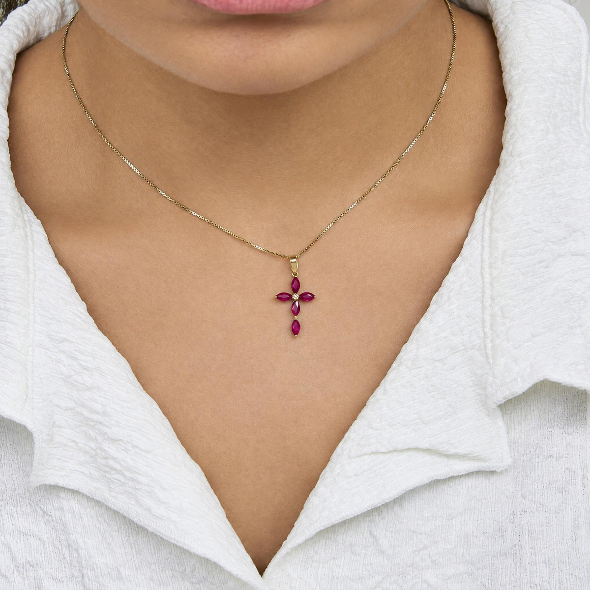 Close-up front view of a model wearing a yellow metal necklace with a cross-shaped pendant featuring marquise red gemstones and a single round white gemstone in the center.