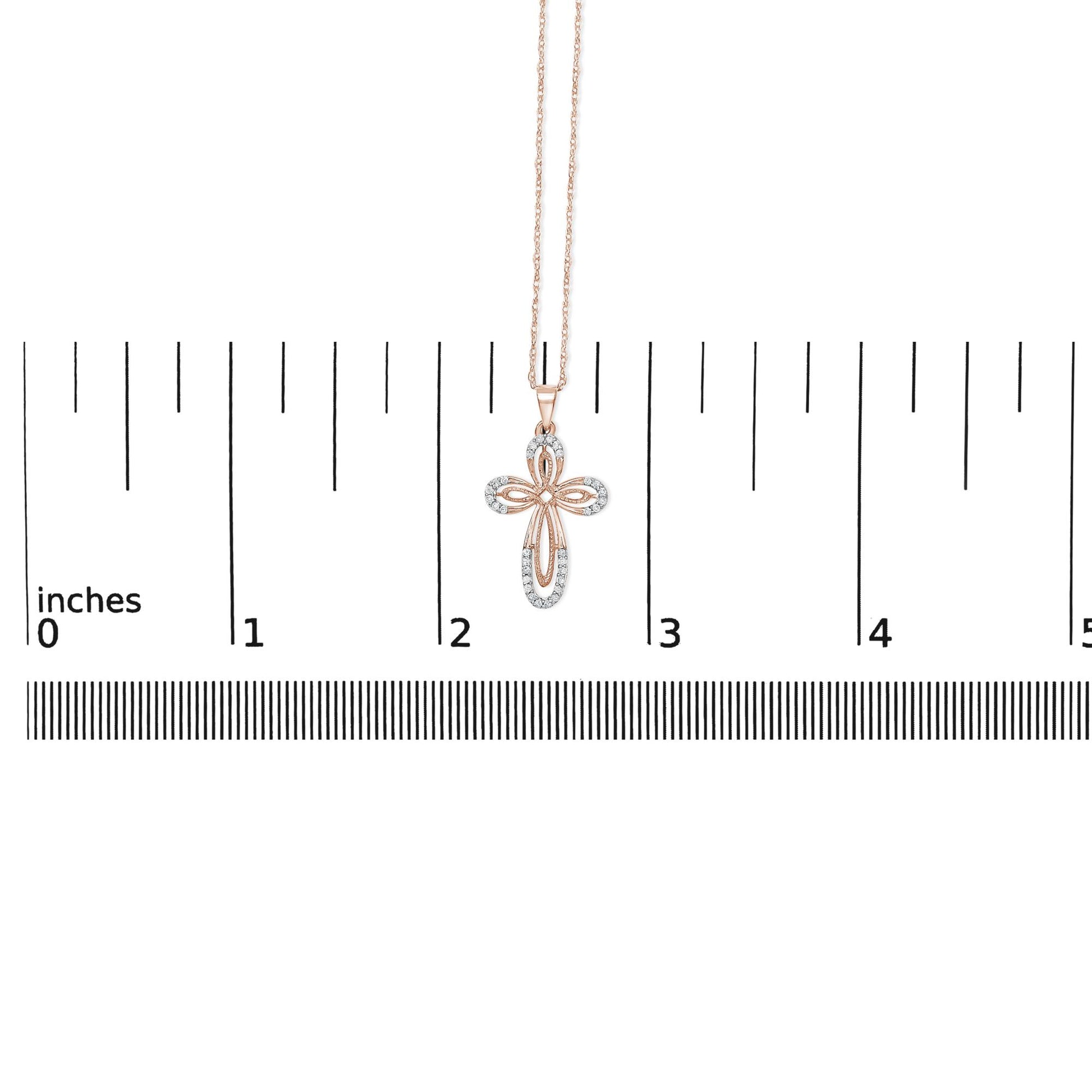 Rose-colored necklace with a cross-shaped pendant featuring round white gemstones in prong settings, shown hanging against a ruler scale, front view.