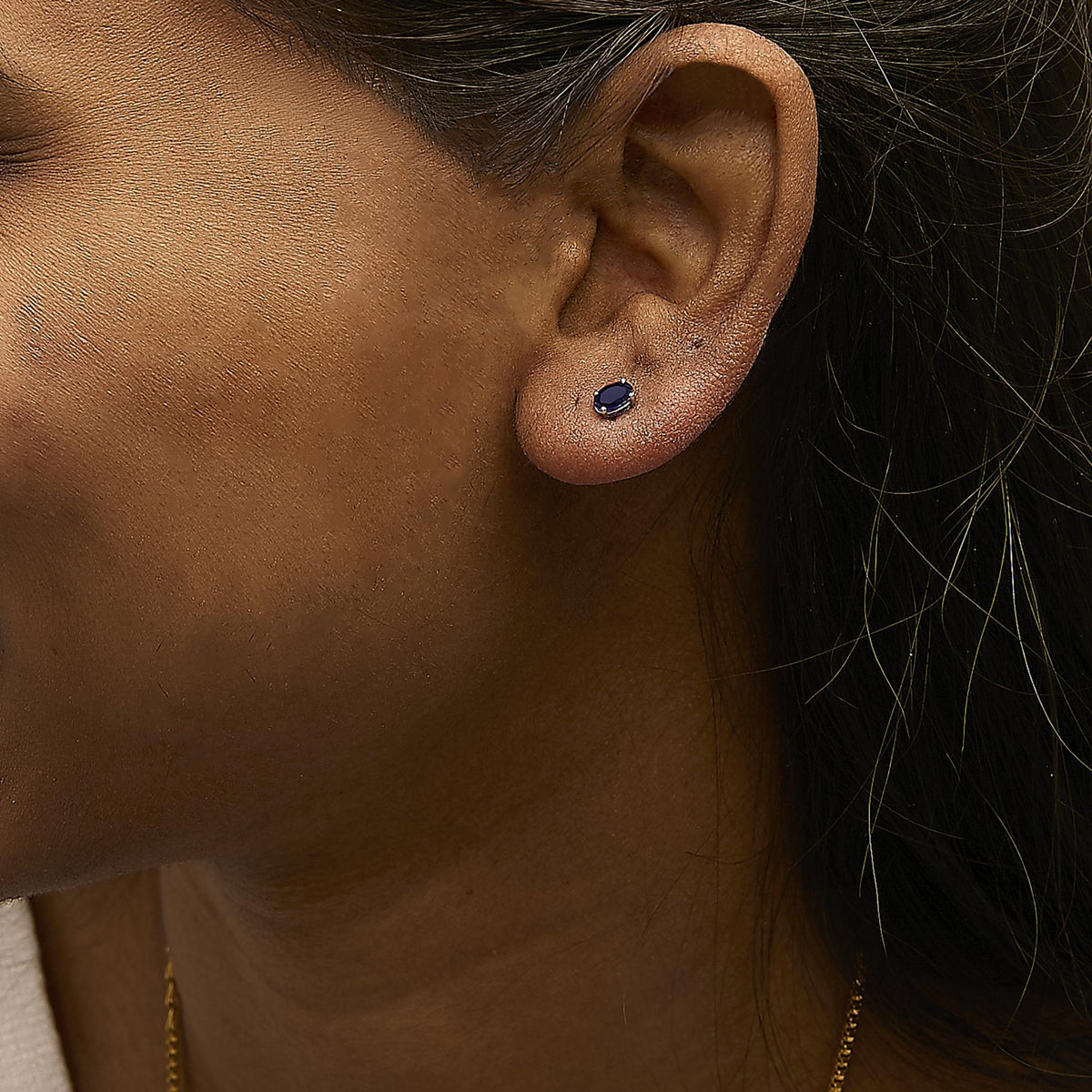 Close-up side view of a model wearing a white metal stud earring featuring an oval-shaped blue gemstone in a four-prong setting.