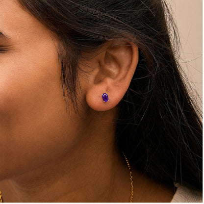 Close-up side view of a model wearing a white metal earring with an oval purple gemstone in a four-prong setting.