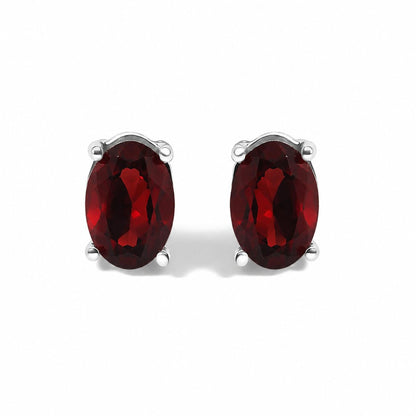 Close-up front view of a pair of white metal earrings each featuring an oval-shaped red gemstone in a four-prong setting, shown on a white background.