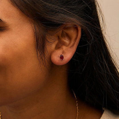 Close-up side view of a model wearing white metal stud earrings featuring oval-shaped red gemstones in a four-prong setting.