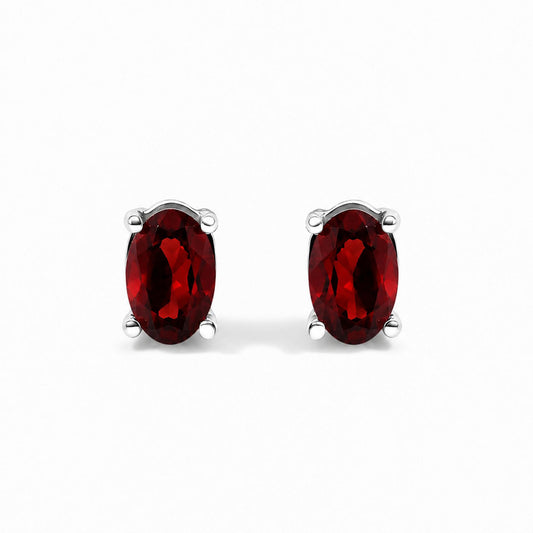 Close-up front view of a pair of white metal stud earrings featuring oval-shaped red gemstones in four-prong settings against a white background.