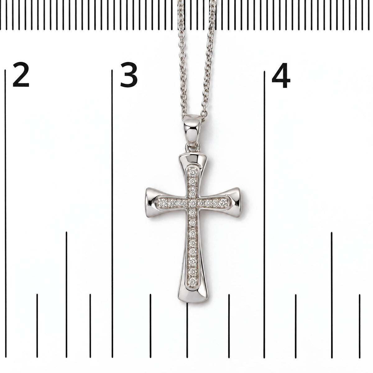 Close-up front view of a white metal cross-shaped necklace pendant with round white gemstones in prong settings on a chain, shown on a measurement scale.