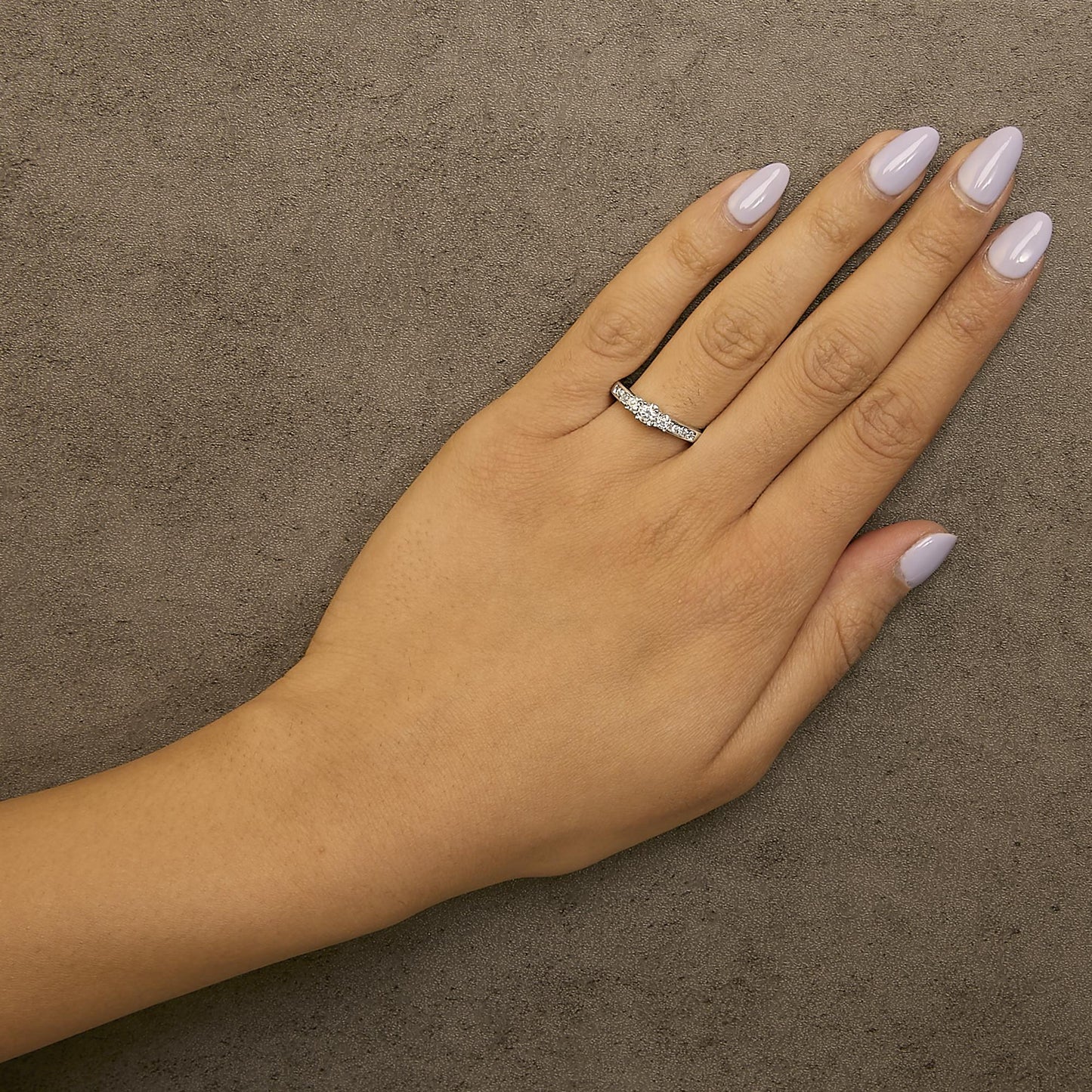 White metal ring with a central round white gemstone in a four-prong setting, flanked by two round white gemstones in four-prong settings, and six small round white gemstones set in a channel, shown worn on a model's hand from a top-down angle.