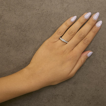 White metal ring with a central round white gemstone in a four-prong setting, flanked by two round white gemstones in four-prong settings, and six small round white gemstones set in a channel, shown worn on a model's hand from a top-down angle.
