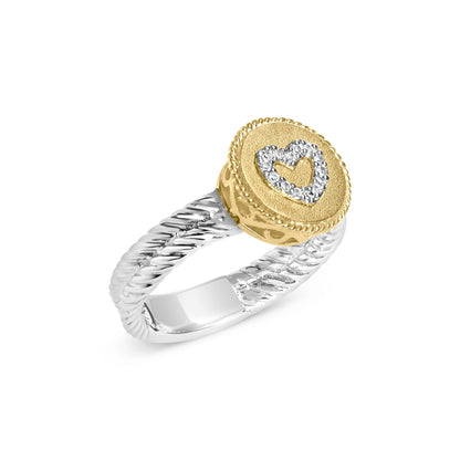 Angled close-up of a two-tone ring with a twisted white band and a textured yellow circular face featuring a heart shape outlined with white gemstones in a shared prong setting.