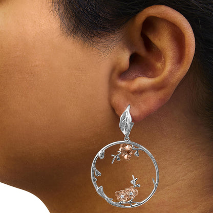 Close-up side view of a white and rose metal hoop earring on a model's ear, featuring floral accents with white gemstones.