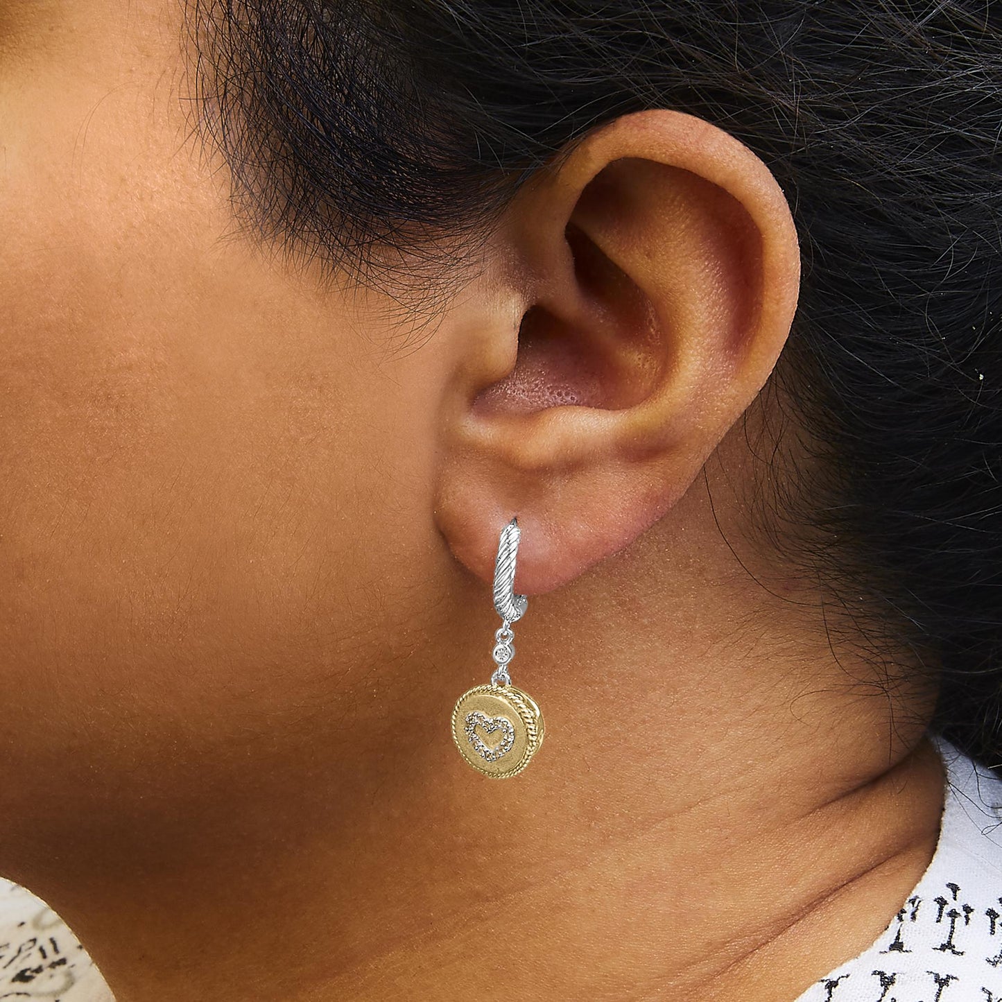 Close-up side view of a model wearing dangle earrings with a twisted white metal hoop, two small white gemstones, and a round yellow metal charm featuring a heart outline of white gemstones.