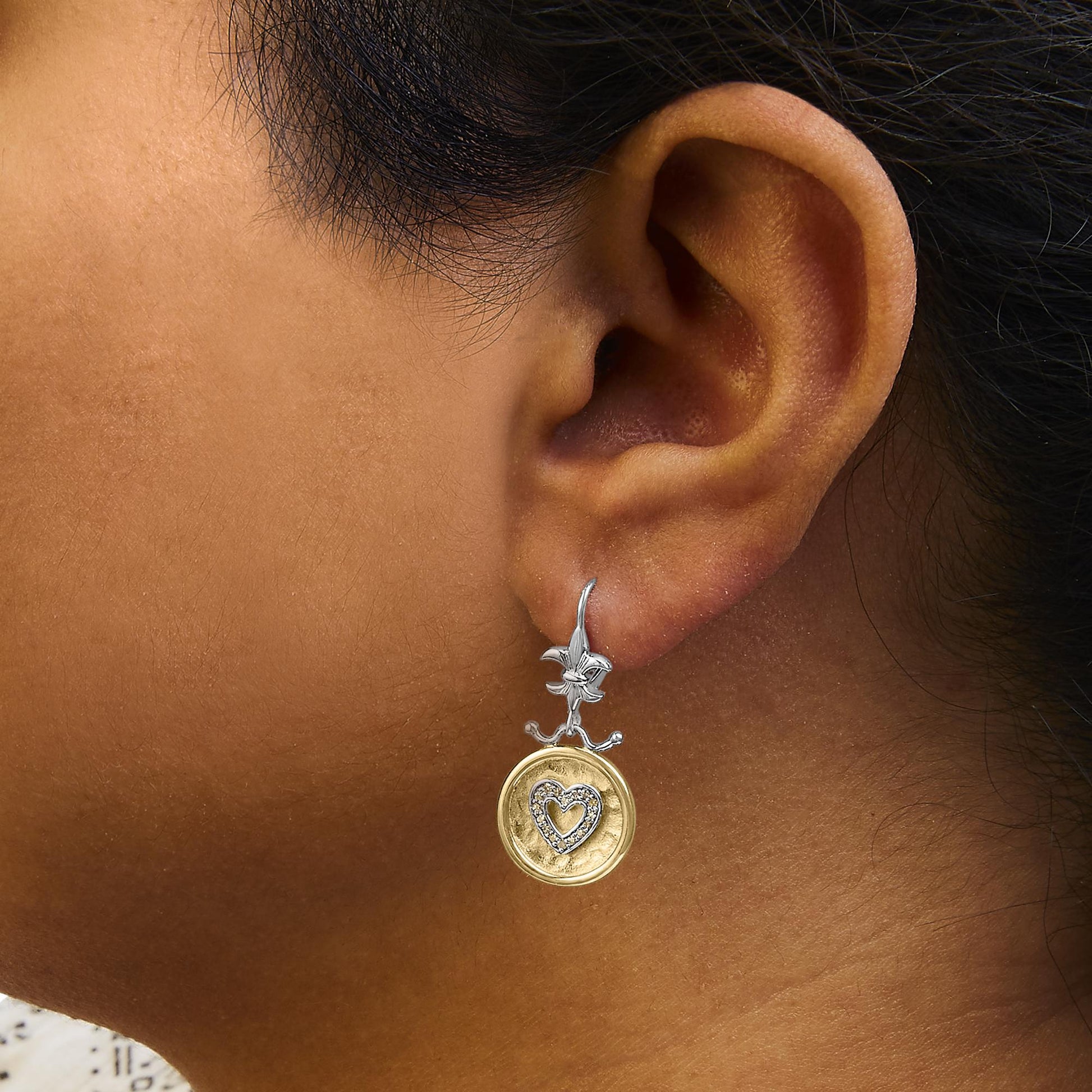 Close-up side view of a model wearing a dangle earring with a white metal flower at the top, a yellow circular pendant, and a heart shape with white gemstones in the center.