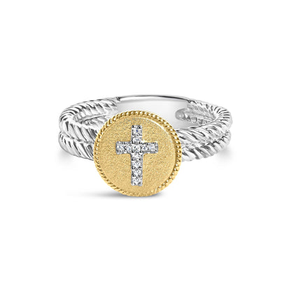 Front angled view of a two-tone ring with a twisted white band and a textured yellow circular top featuring a cross made of round white gemstones in shared prong setting.