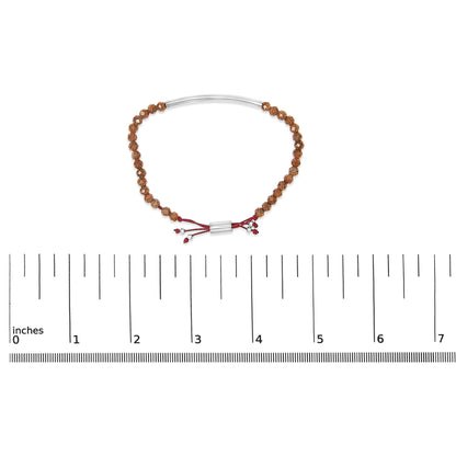 Bracelet with brown textured beads and a white curved bar, red adjustable cord with red and white gemstones, shown on a scale from a top-down angle.