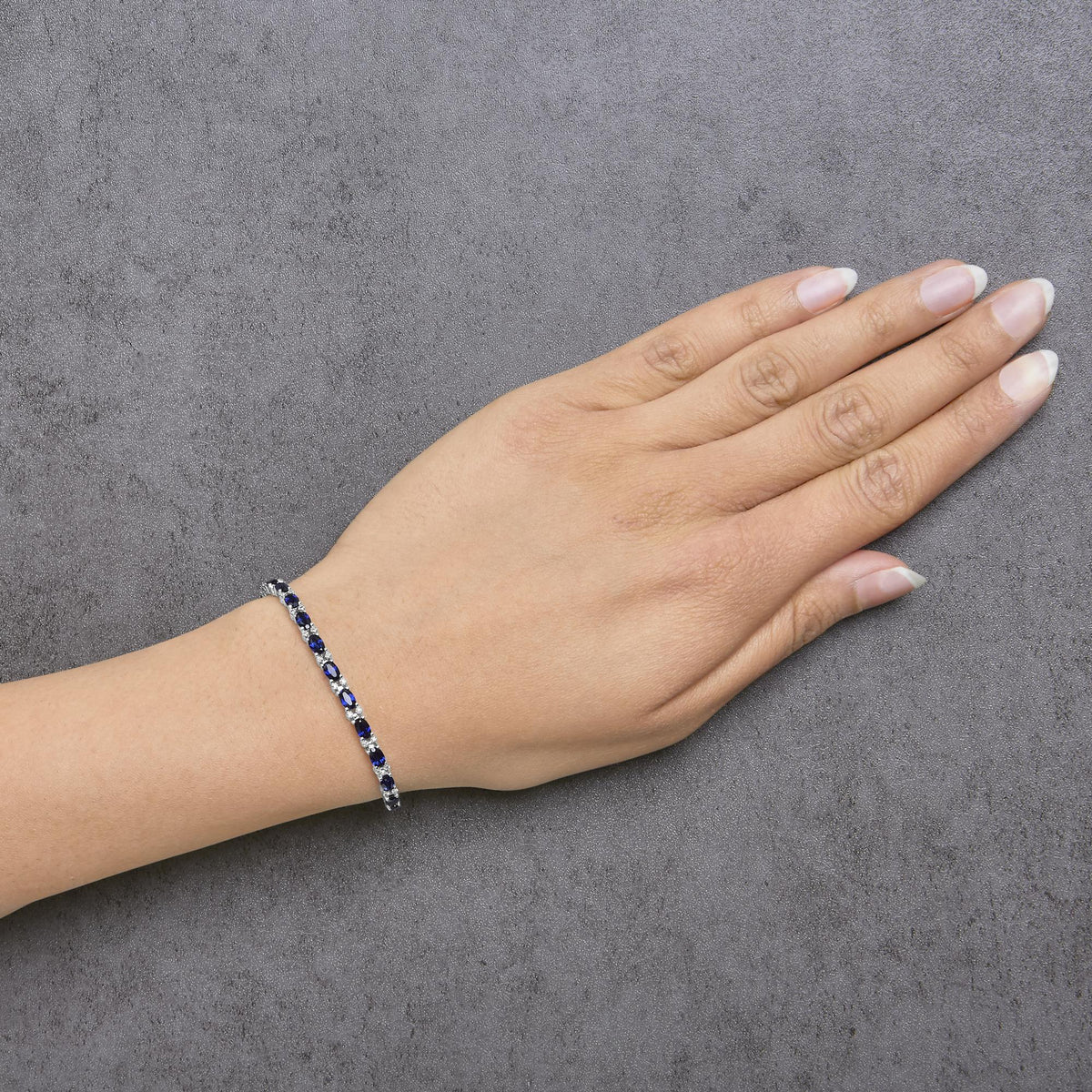 White metal bracelet with alternating oval blue gemstones and round white gemstones in prong settings, shown worn on a model's wrist from a top angle.