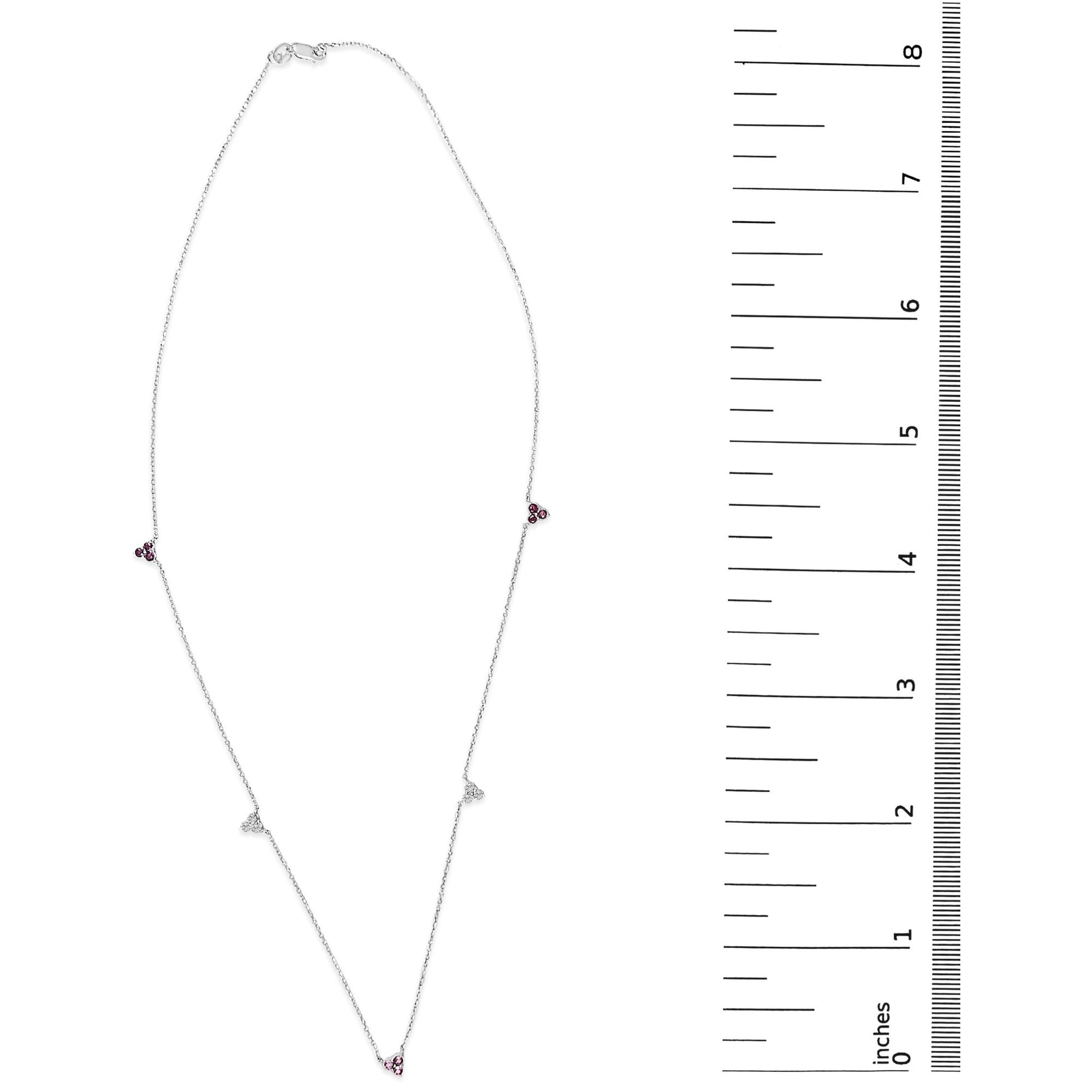 White metal necklace shown on a scale, featuring small triangular clusters of red and white round gemstones in prong settings spaced along a delicate chain. The image is a top-down view.