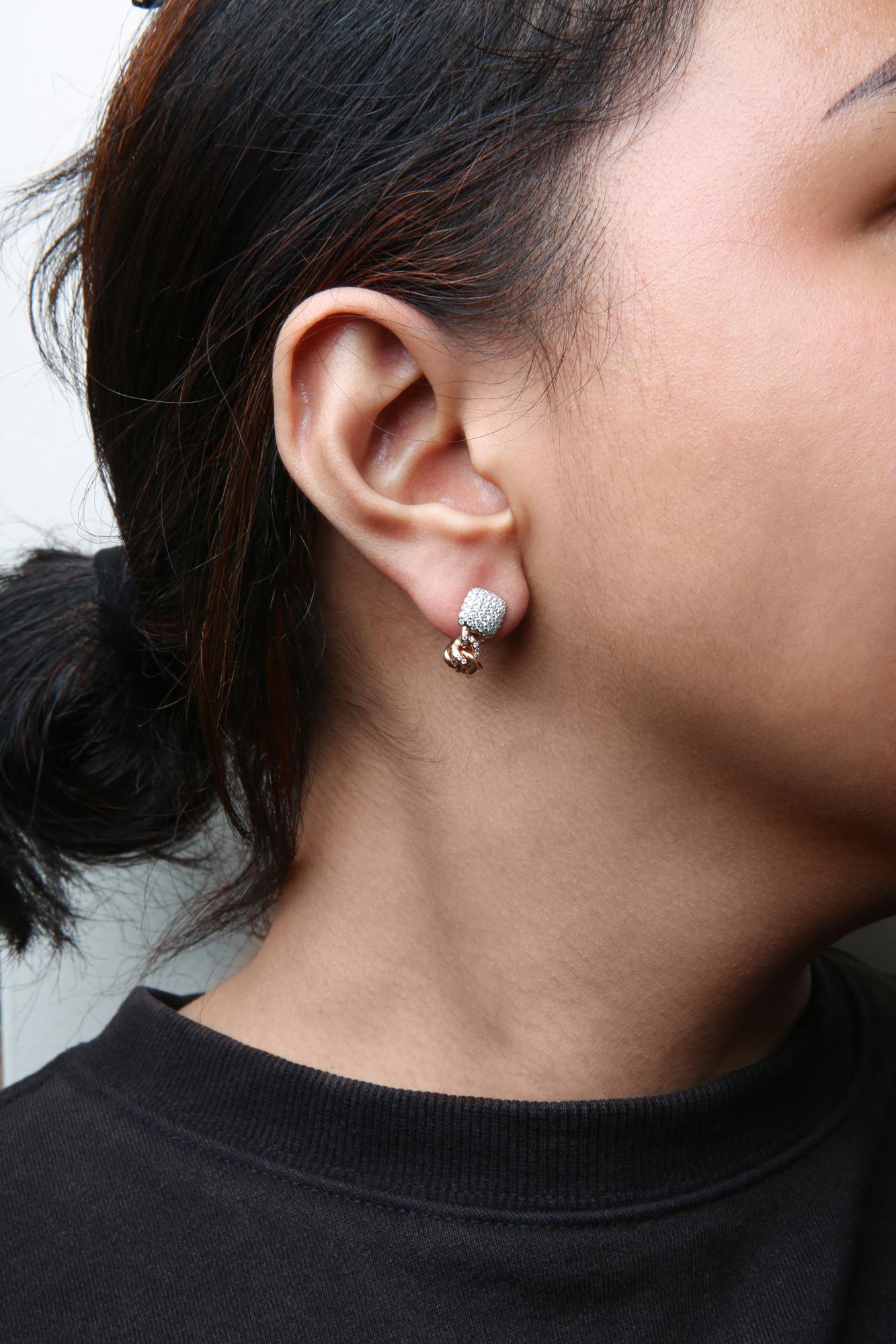 Close-up side view of a rose-colored metal earring on a model's ear, featuring a cluster of prong-set white round gemstones on the front with a small hanging rose-colored element below.