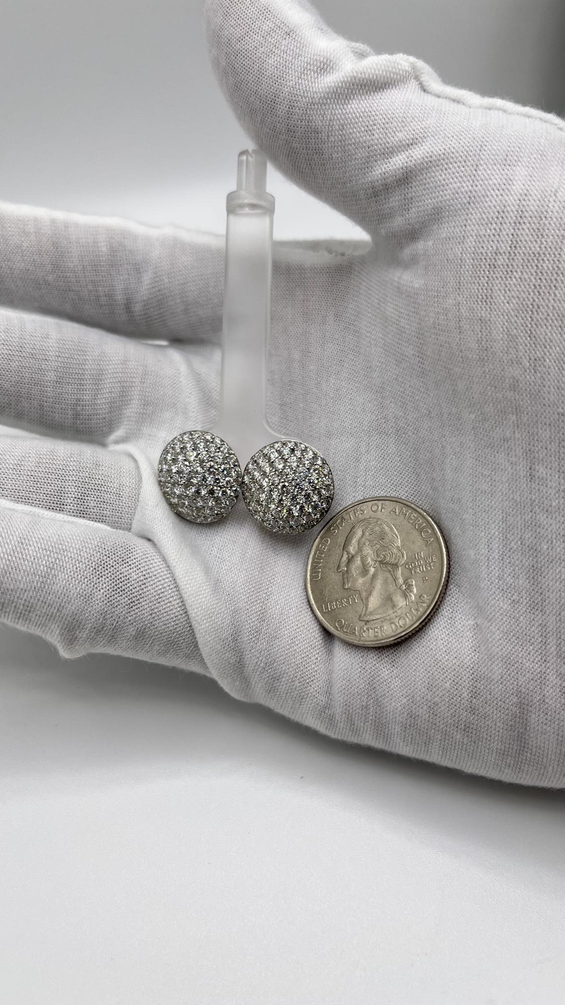 Close-up front angle of a pair of white metal round-shaped shared prong set white gemstone stud earrings held in a white gloved hand next to a quarter coin for size reference.