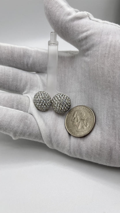 Close-up front angle of a pair of white metal round-shaped shared prong set white gemstone stud earrings held in a white gloved hand next to a quarter coin for size reference.