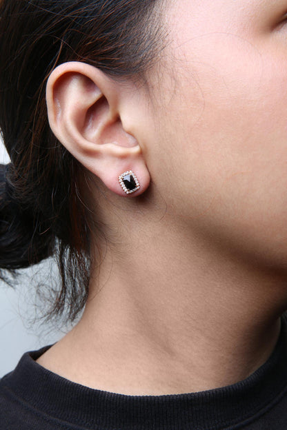 Close-up side view of a model wearing square cushion-shaped black gemstone stud earrings surrounded by small round white gemstones, set in white and rose metal.