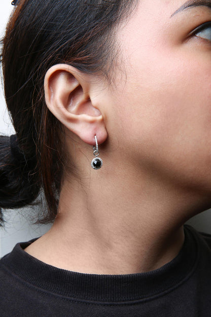 Close-up side view of a model wearing white metal hoop earrings with small round white gemstones set along the hoop and a black round gemstone surrounded by small white gemstones hanging from each hoop.
