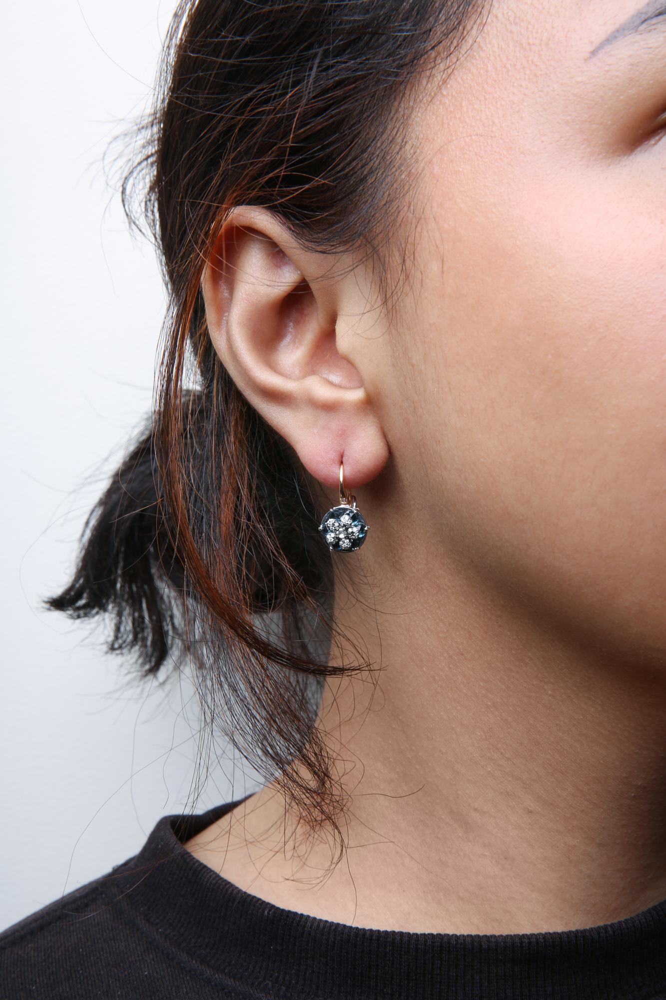 Close-up side view of a woman's ear wearing a white and rose metal earring featuring a central blue gemstone surrounded by multiple small white gemstones.