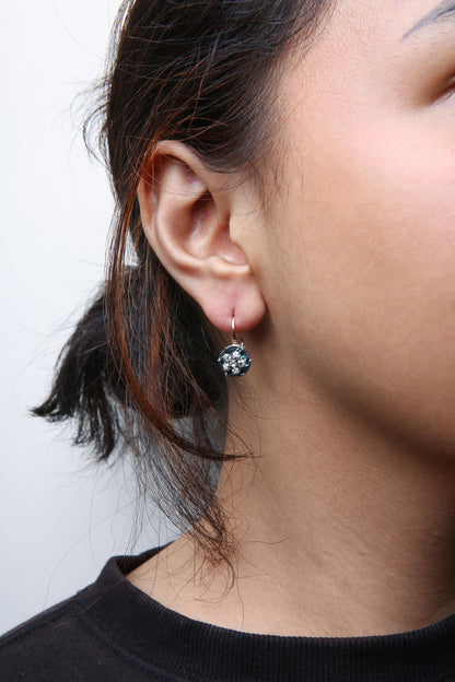 Close-up side view of a woman's ear wearing a white and rose metal earring featuring a central blue gemstone surrounded by multiple small white gemstones.