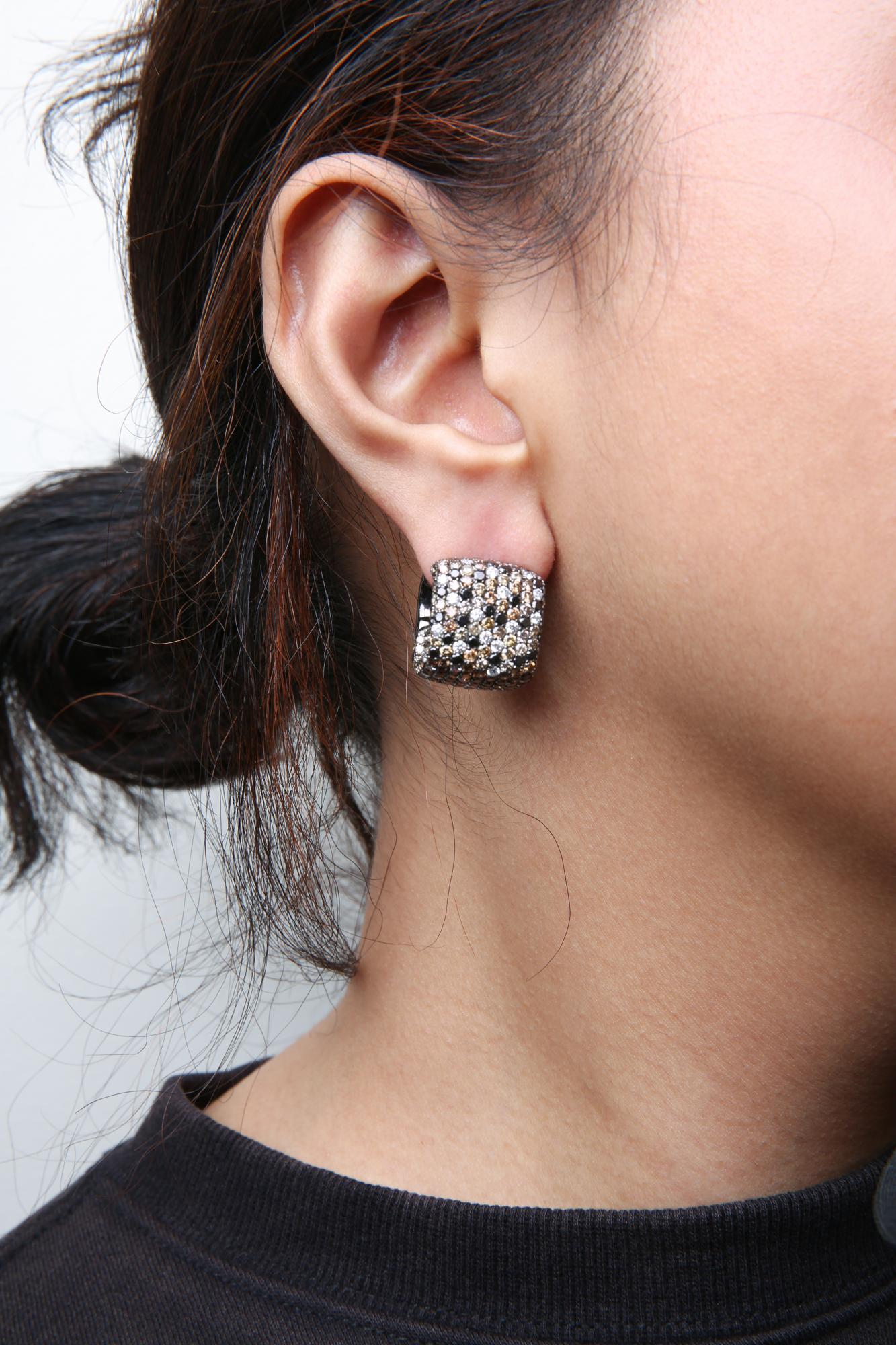 Close-up side view of a woman's ear wearing a white and black metal earring encrusted with brown, black, and white round pave-set gemstones.