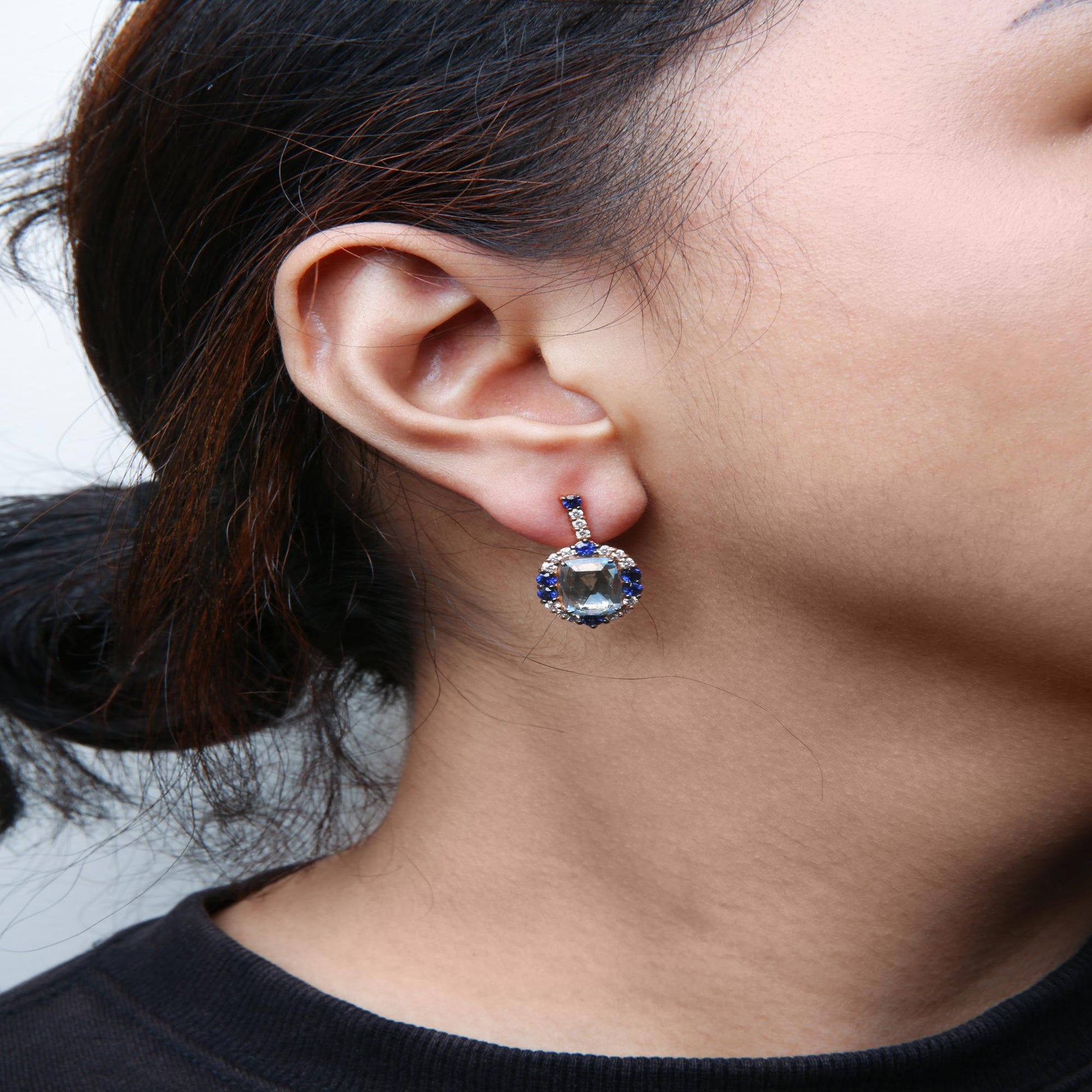 Close-up side view of a model wearing drop earrings with white and rose metal, featuring a cushion-shaped blue gemstone surrounded by round blue gemstones and white gemstones in prong settings.