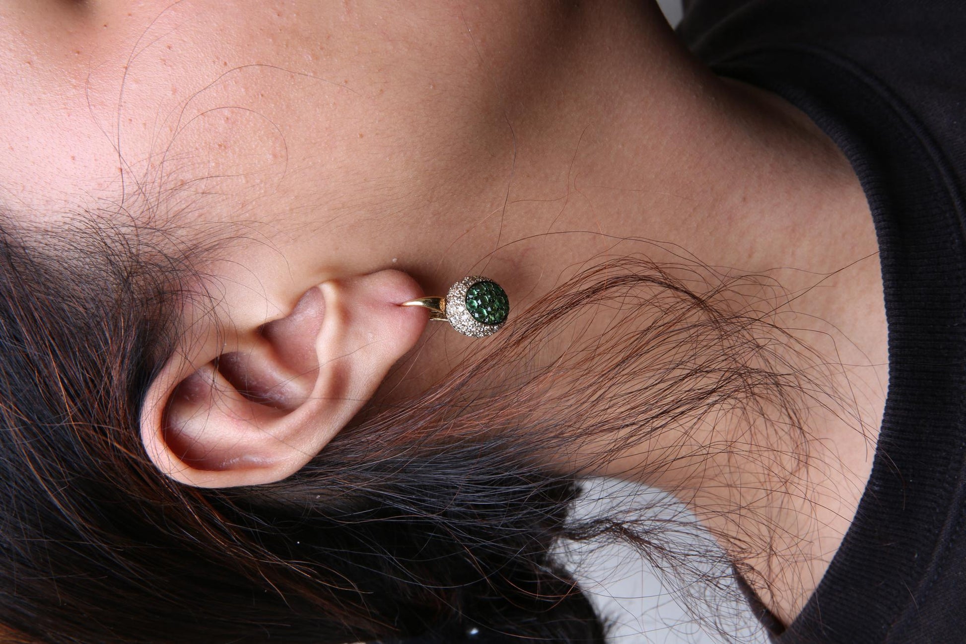 Close-up angled shot of a model wearing a yellow and black metal earring featuring a cluster of round green gemstones surrounded by a pave setting of round brown gemstones.