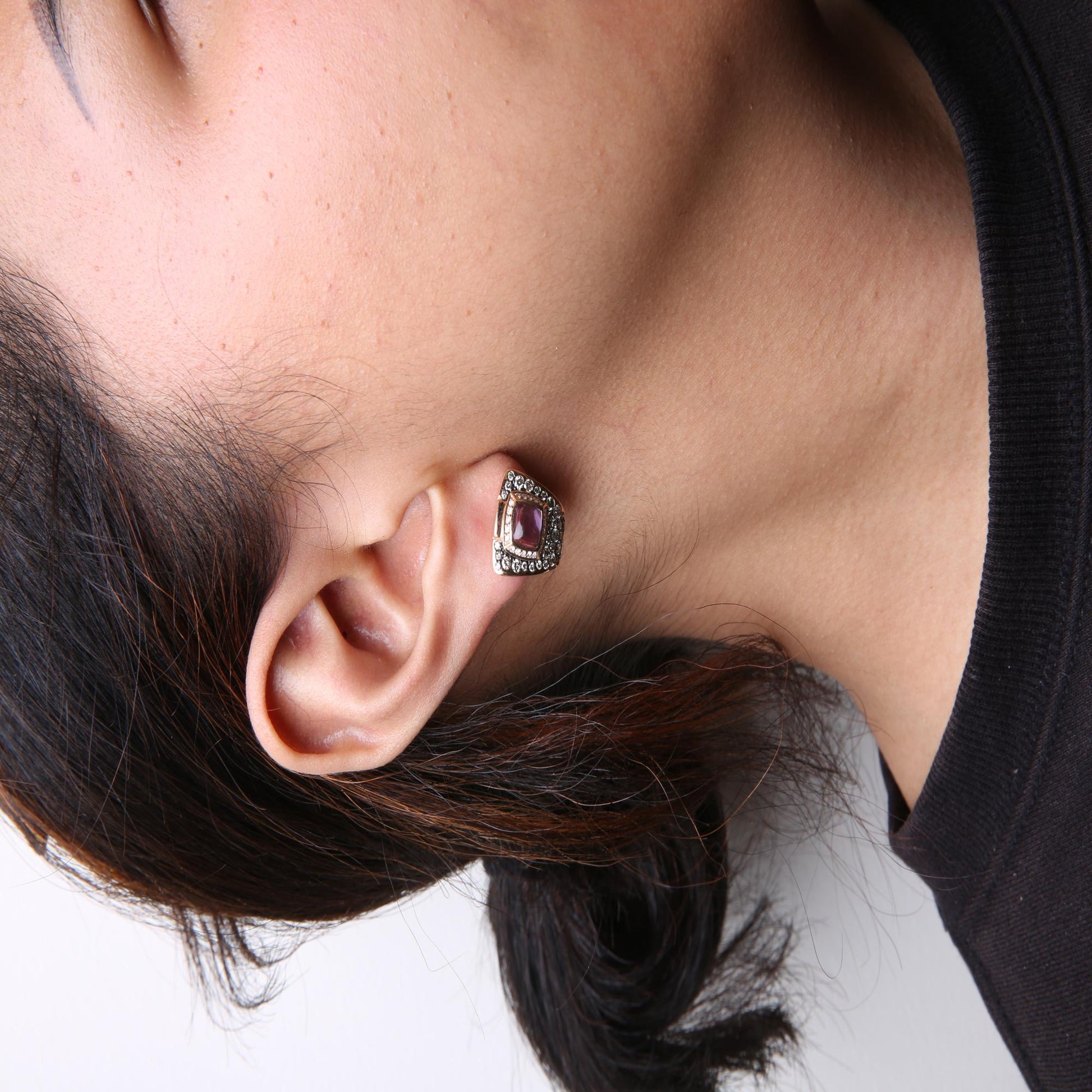 Close-up side view of a model wearing rose-colored earrings with a cushion-shaped purple gemstone at the center, surrounded by brown and white round gemstones in pave and channel settings.