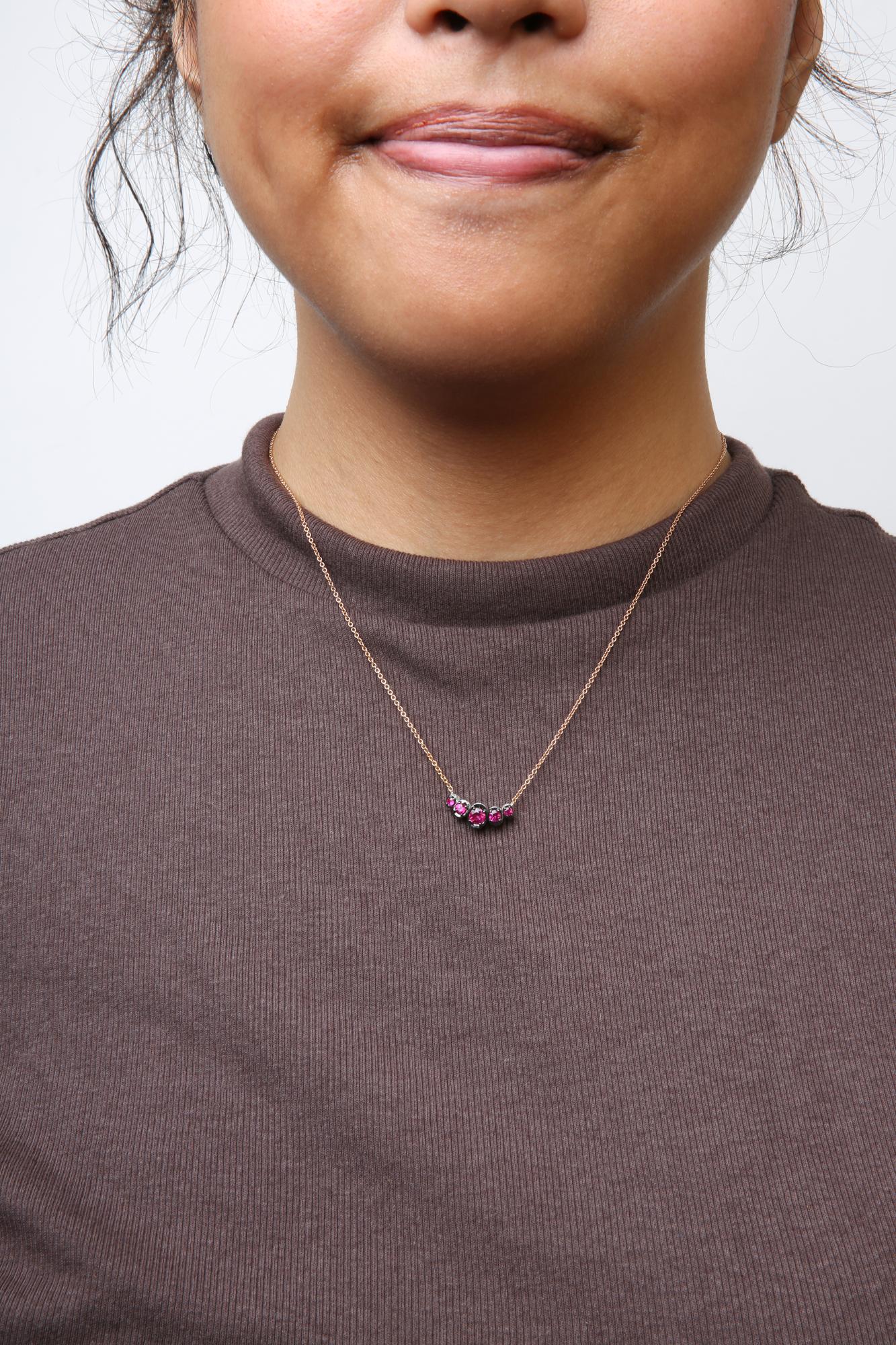 Close-up front view of a model wearing a rose metal necklace with a curved pendant featuring round red gemstones in prong settings and small white gemstones in pave settings.