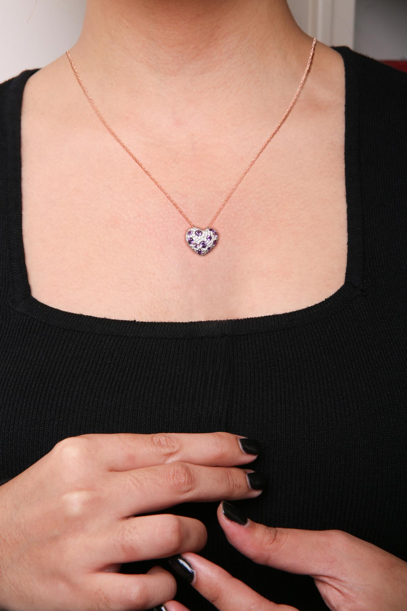 Close-up front view of a rose metal necklace on a model featuring a heart-shaped pendant adorned with round white and purple gemstones in prong settings, the model wears a black top and has black nail polish.