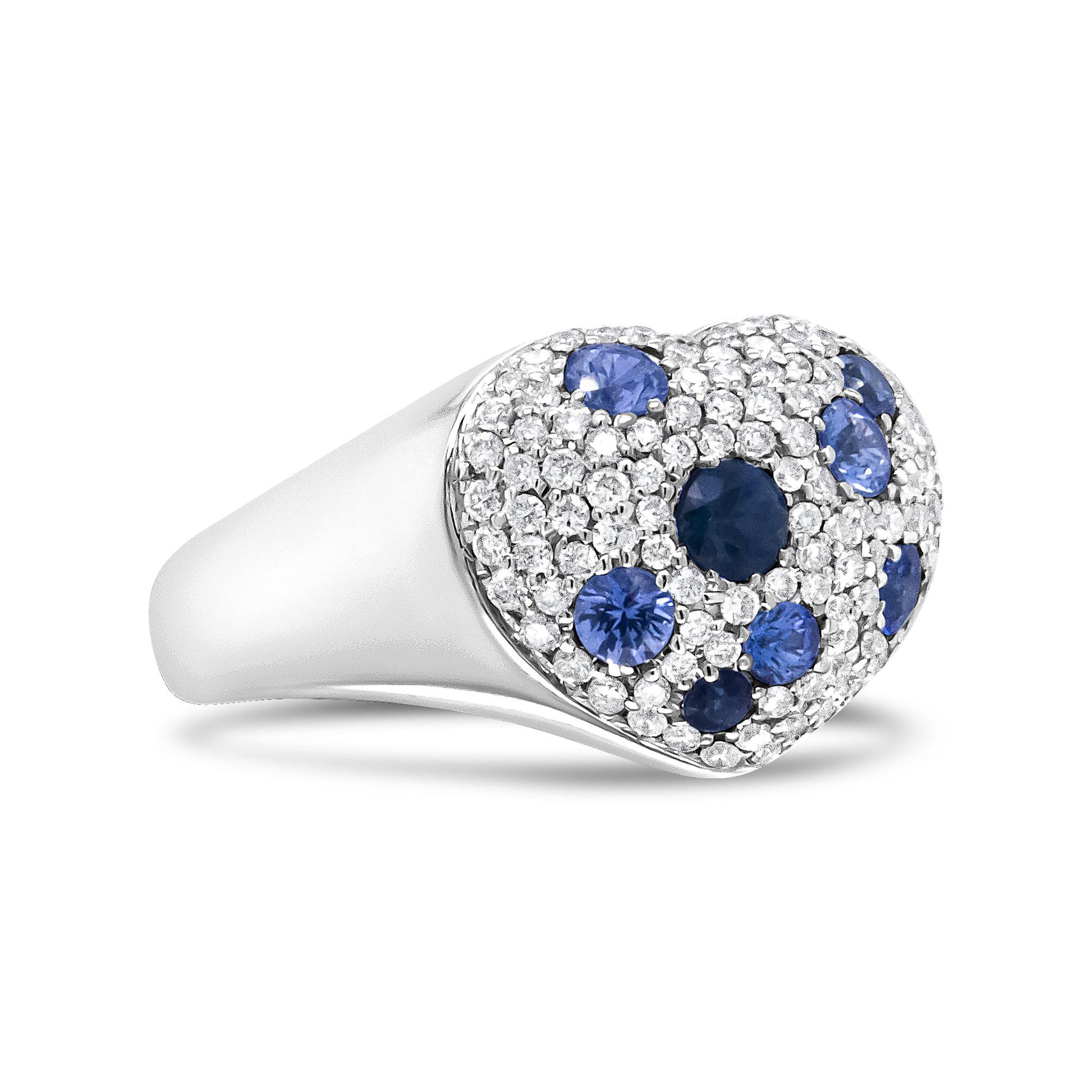 Close-up side angle of a white metal ring with a heart-shaped top featuring a clustered pave setting of round white gemstones and scattered round blue gemstones.