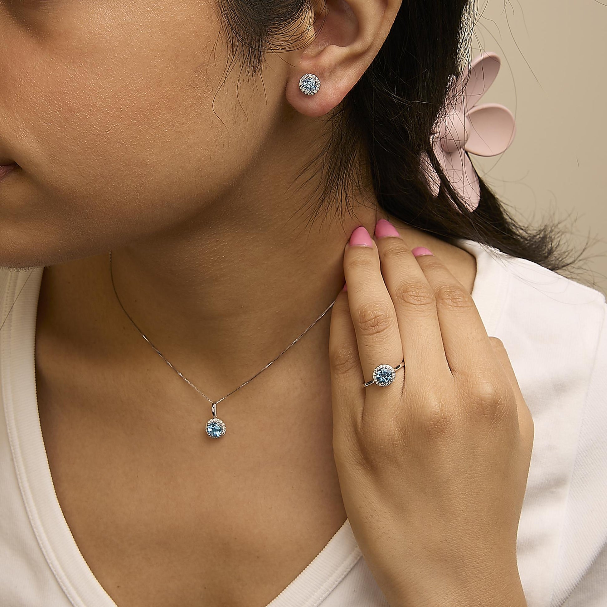 Close-up side view of a model wearing a white metal jewelry set including round blue gemstone stud earrings, a matching pendant necklace, and a ring, all surrounded by small white gemstones.