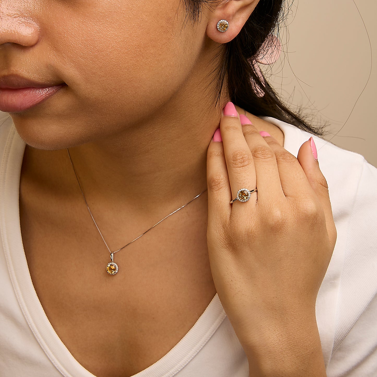 Close-up front angle of a model wearing a jewelry set including round orange gemstone stud earrings, a matching pendant necklace, and a ring, all surrounded by small white gemstones with shared prong settings on white metal.