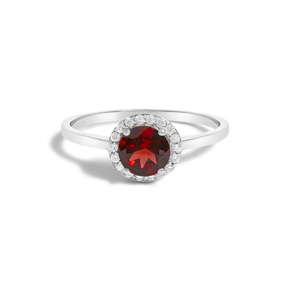 Close-up front angle of a white metal ring featuring a round red gemstone in a four-prong setting, surrounded by small round white gemstones in a shared prong setting.