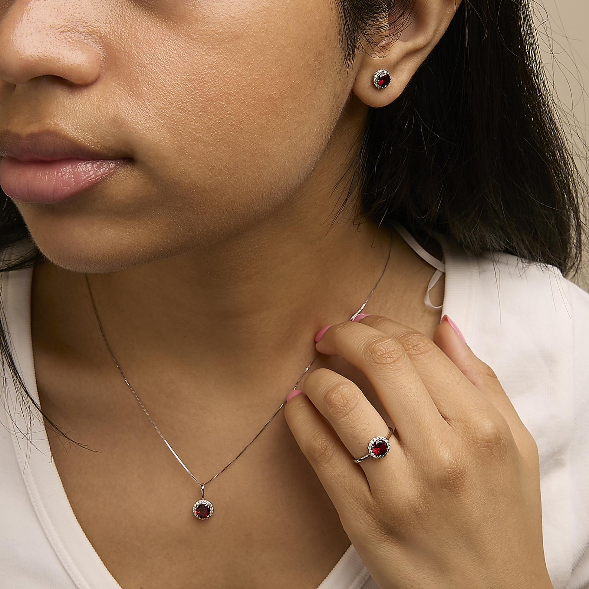 Set of jewelry shown worn on a model, including round red gemstone stud earrings, a matching pendant necklace, and a ring, all surrounded by small white gemstones, shot from a close-up angle.