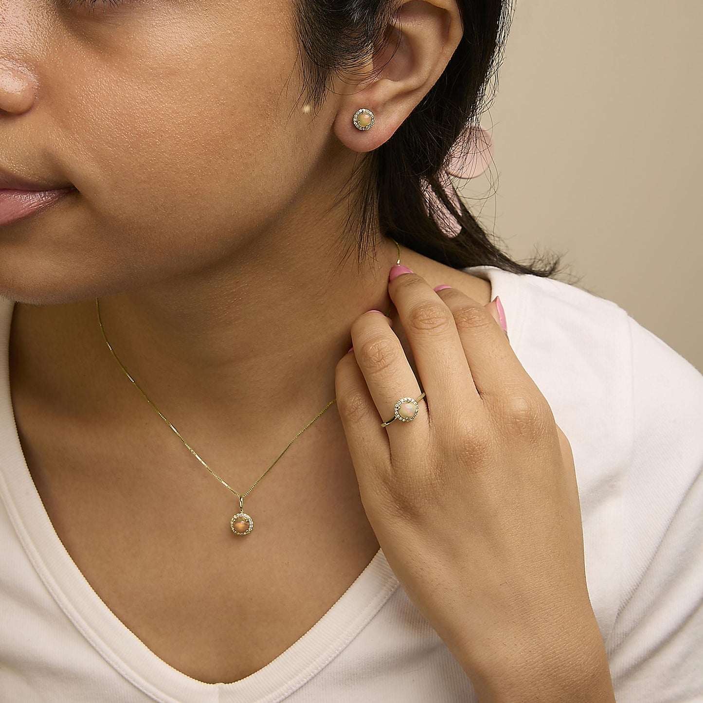 Close-up angled shot of a model wearing a jewelry set including round multi-color gemstone stud earrings, a matching pendant necklace, and a ring. Each piece features a round gemstone centered and surrounded by small white gemstones in shared prong settings on yellow metal.