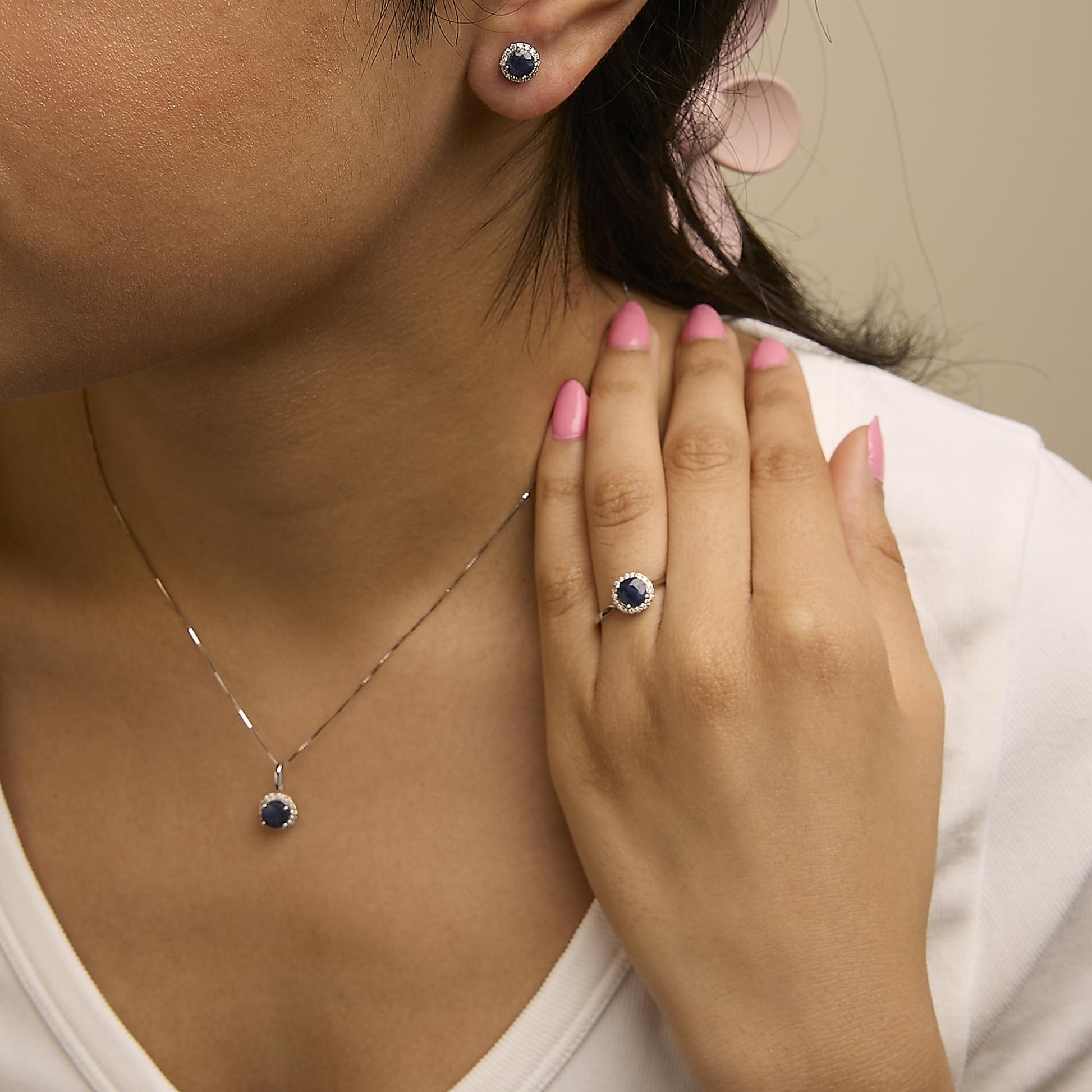 Close-up angled shot of a model wearing a jewelry set featuring a necklace pendant, ring, and stud earrings. Each piece has round blue gemstones surrounded by small white gemstones set in white metal.