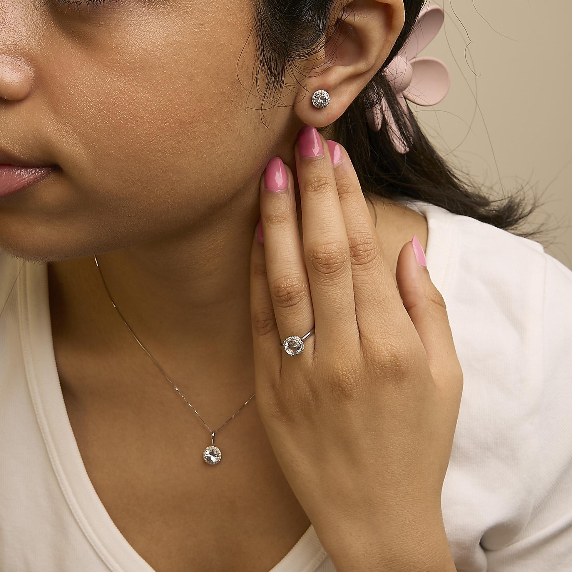 Set of white metal jewelry including round-shaped white gemstones surrounded by smaller round white gemstones in shared prong settings, shown worn on a model’s ear as earrings, finger as a ring, and neck as a pendant, front-facing angle.